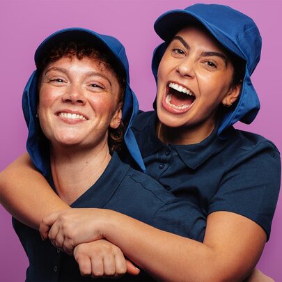 Two young people wearing blue sunhats and shirts, arm in arm in front of a purple background.