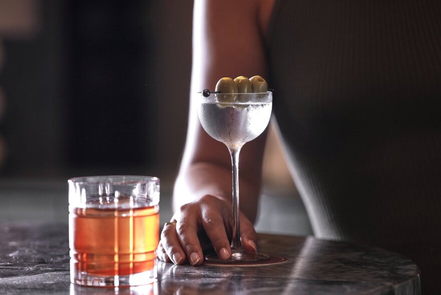 A woman's hand resting on the bottom of a tall  martini glass, alongside a shortball glass with a whiskey cocktail.