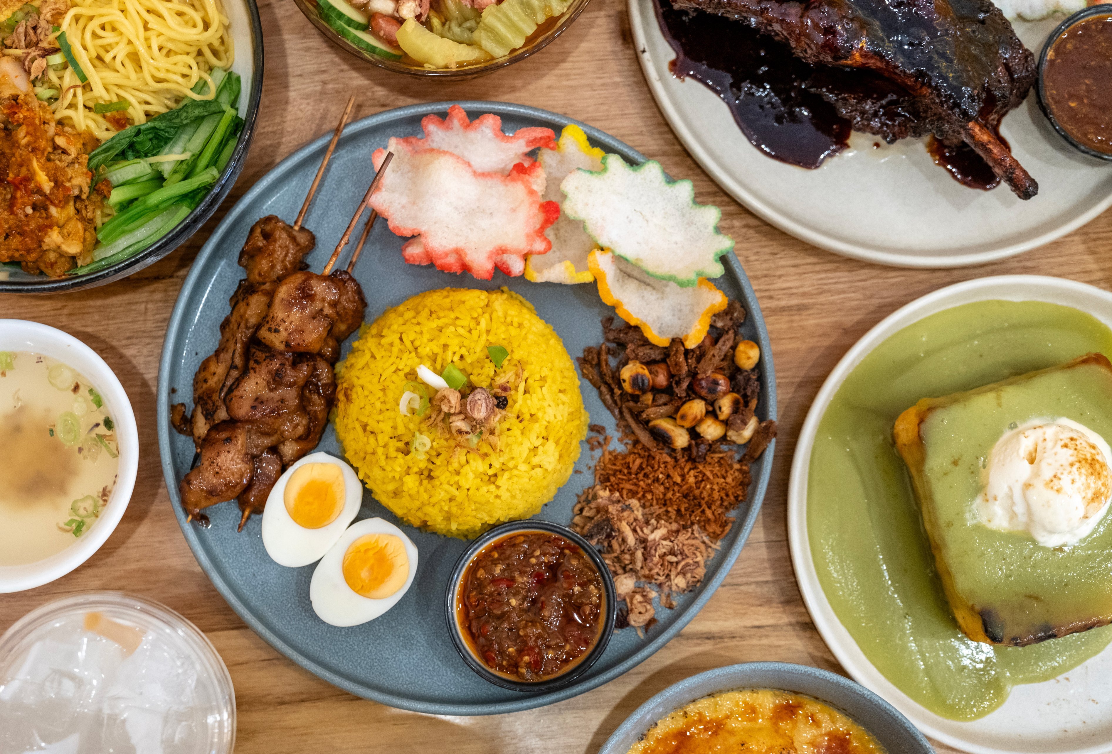 An assortment of Indonesian dishes including noodles, beef ribs, chicken satay skewers, yellow rice and pandan toast.