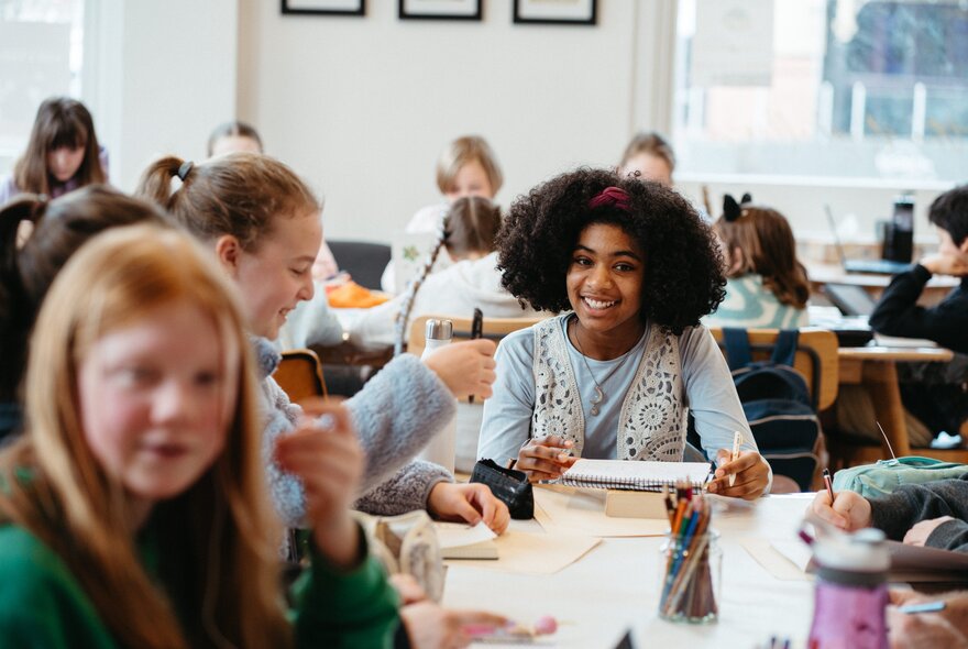 A girl with curly black hair smiling at a table in a classroom setting with kids at long tables. 