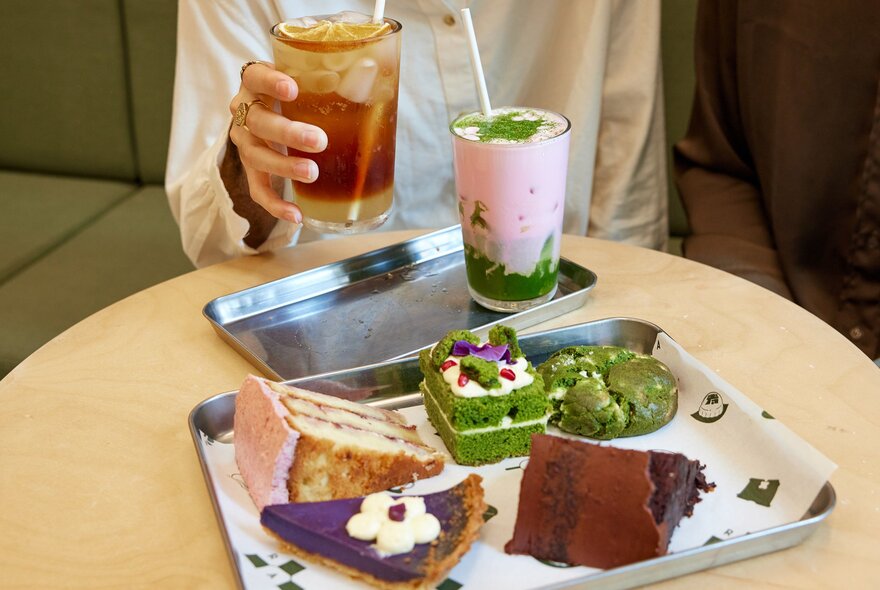 A person is sipping on an iced tea in a cafe. In front of them is a ray of cakes and cookies.