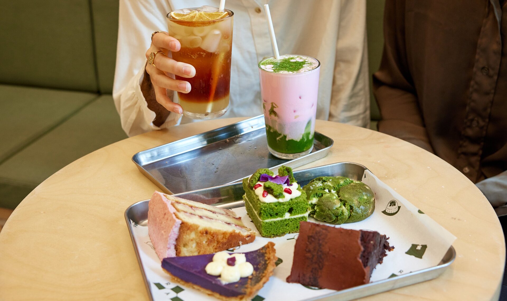 A person is sipping on an iced tea in a cafe. In front of them is a ray of cakes and cookies. 