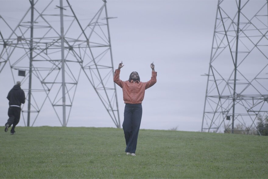 An open grassy field with large, metallic electricity pylons towering in the background under an overcast, grey sky and two people in the area.