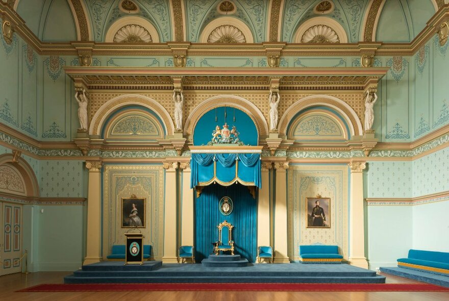 A stately room at Government House Victoria, with a blue velvet throne style chair on a low rise stage, blue velvet bench seating on either side and architectural wall trims of arches and columns.