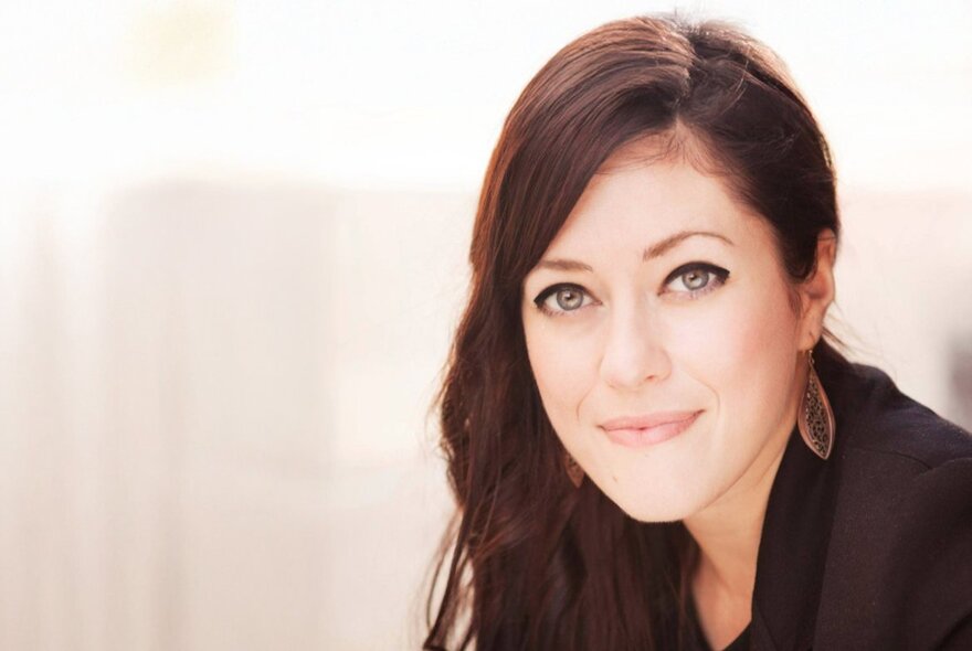 Author Callie Hart, a smiling young woman with long brown hair and a dark top, wearing drop earrings and posed against a light coloured backdrop.