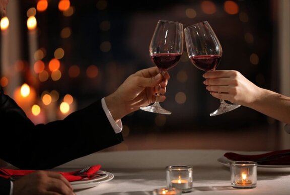 Two arms clinking glasses of red wine over small tealights with other candles in the background. 