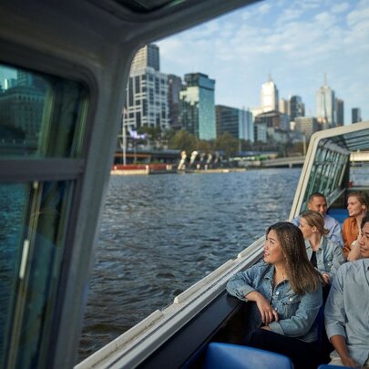 People seated in a river boat with outside seating, passing city buildings.