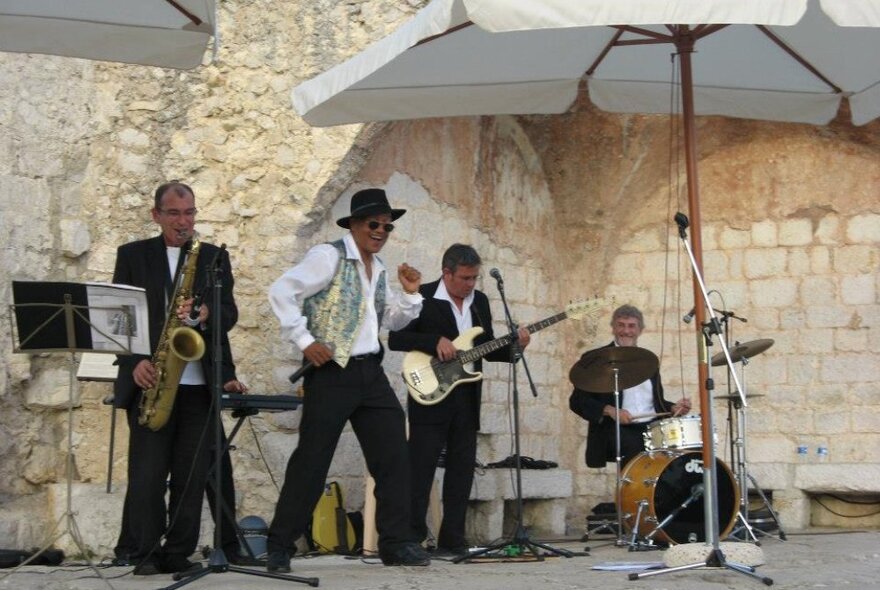 Jazz band performing under umbrellas on a stage set against crumbling white stone.