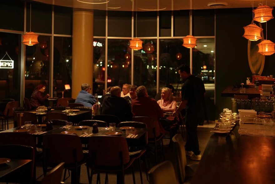 A warm, dimly-lit dining space with people at a long table and glowing lanterns