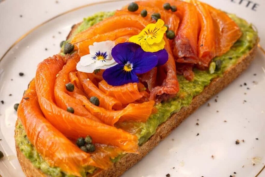 Smoked salmon on a slice of bread on a plate, garnished with colourful pansies and capers.