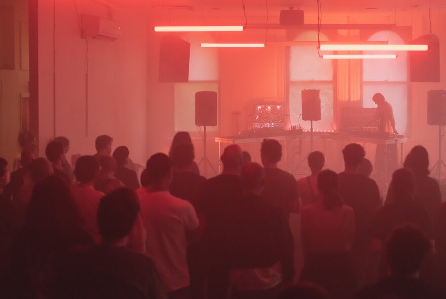 A red filtered light over a room with a small audience watching a DJ. 
