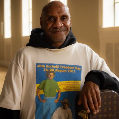 A smiling Amos Roach, wearing a white slogan t-shirt and standing in a large empty room.