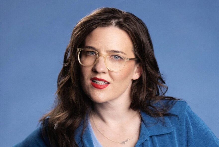 Comedian Rose Callaghan, with straight brown hair, smiling slightly and looking at the viewer, wearing a blue top and glasses.