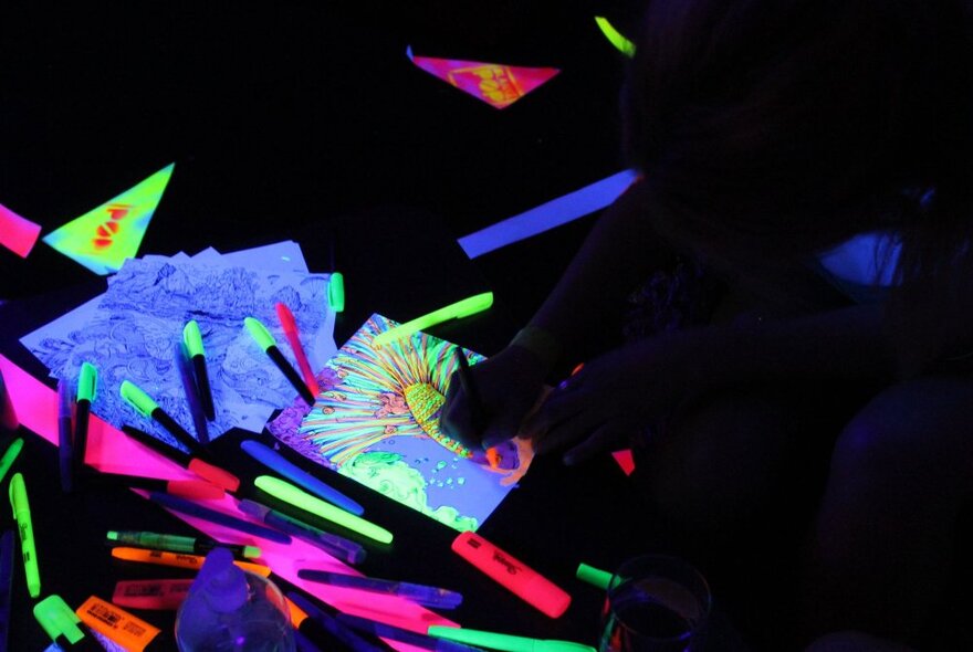 A collection of neon glow-in-the-dark pens and textas alongside colourful drawings on paper in a darkened space.