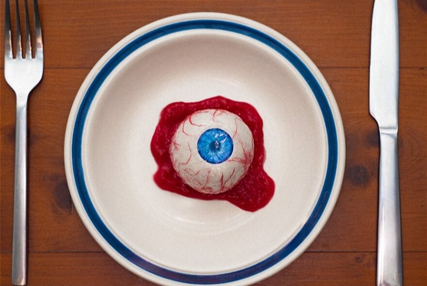 A large, bloodshot eyeball resting in a small pool of blood in the middle of a white dinner plate, with cutlery set beside it.