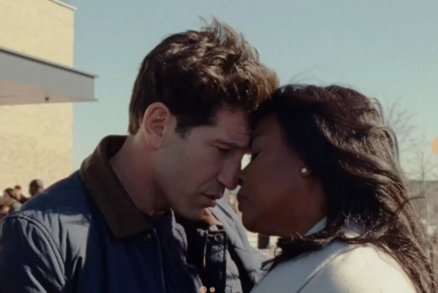 Movie still of a man and woman resting foreheads together outside with blue sky.