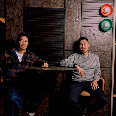 Chefs Cameron Tay and Ian Ho seated at a high table, causally leaning back against a dark wall. 