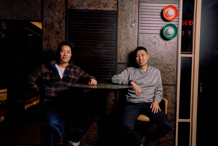 Chefs Cameron Tay and Ian Ho seated at a high table, causally leaning back against a dark wall. 