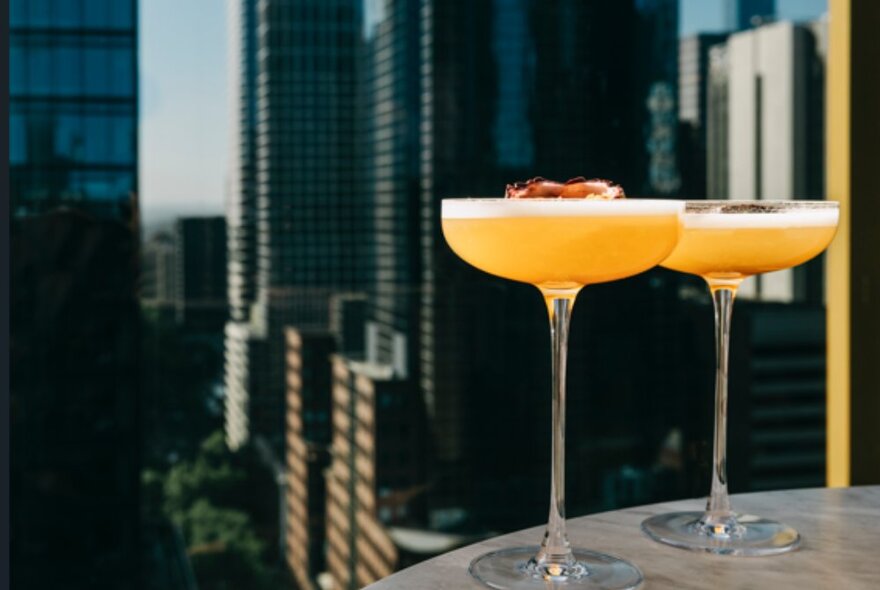 Two long-stemmed cocktail glasses on a round table, with city buildings behind.