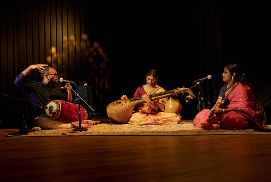 Three musicians seated on stage playing traditional Indian instruments while wearing colourful traditional clothing.