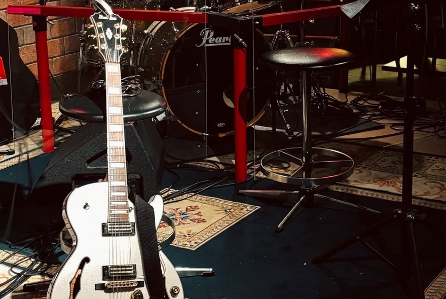 A white electric guitar on a stand, with a drum kit and a black stool behind it.