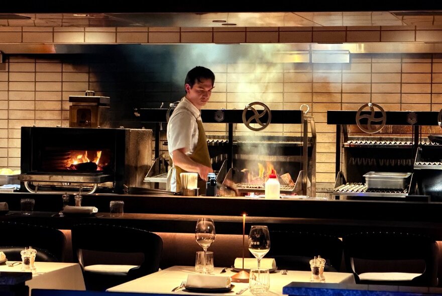 A chef stands behind a bar in an open restaurant kitchen, working beside an active wood-fired grill filled with smoke and flames.