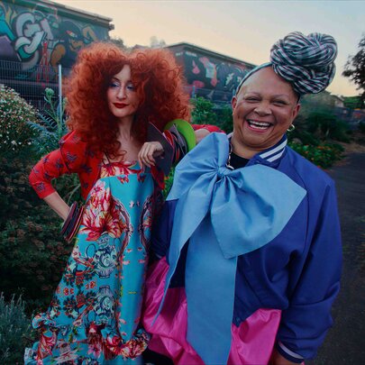 Two models stand smiling outdoors in vibrant, unconventional fashion ensembles.