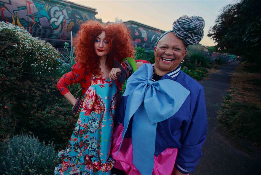 Two models stand smiling outdoors in vibrant, unconventional fashion ensembles.