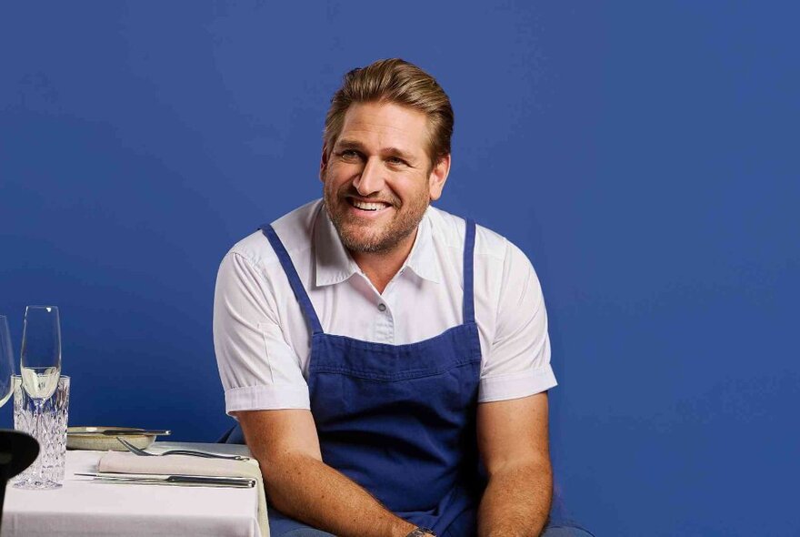 Chef Curtis Stone seated at a white-napery table wearing a blue apron and white shirt.