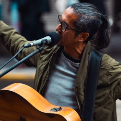 A busker performing in a street with a guitar around his neck and arms outstretched while singing. 