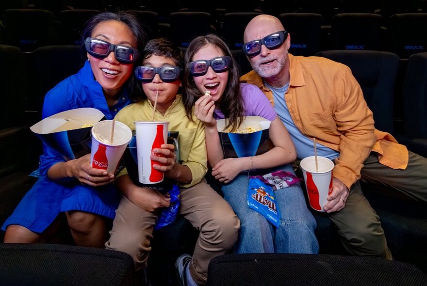 A family of four in a cinema with 3D glasses on, each holding a large popcorn and a large drink.