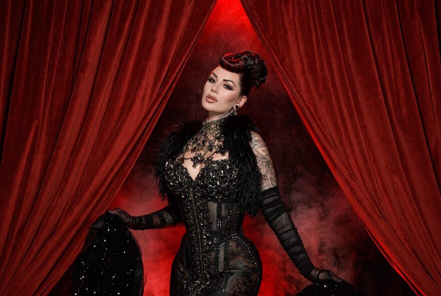 Tattooed burlesque dancer appearing between parted red velvet curtains, wearing black lace.