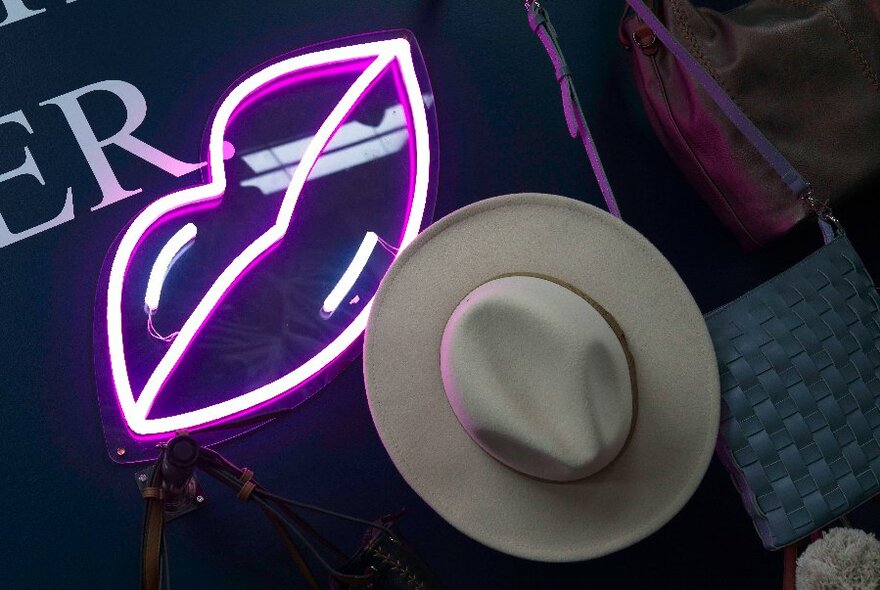 Looking down at a neutral coloured fedora hat next to a neon set of lips. 