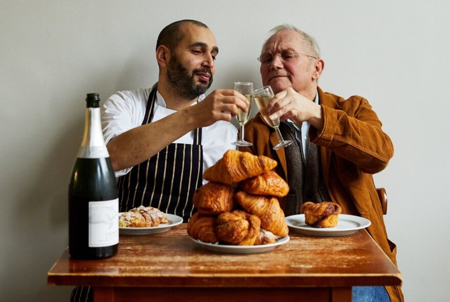 Chefs Trevor Gulliver and Farokh Talati seated at a small table toasting each other with champagne with a pile of croissants in front of them and a bottle. 