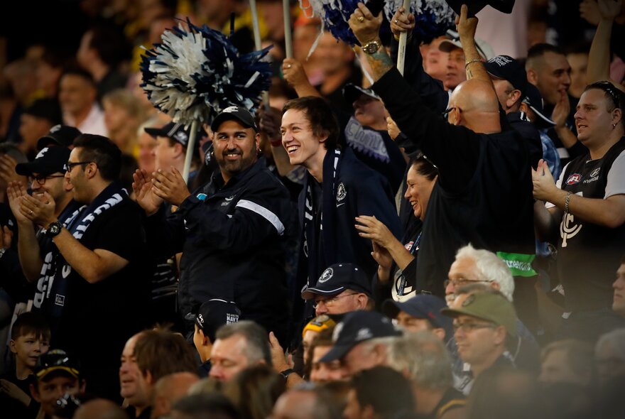 AFL footy fans in the seats at a stadium watching a game and cheering.