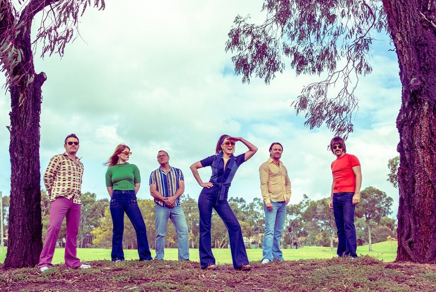 A group of six people wearing 1970s inspired clothing and standing in a large open space between two tree trunks.