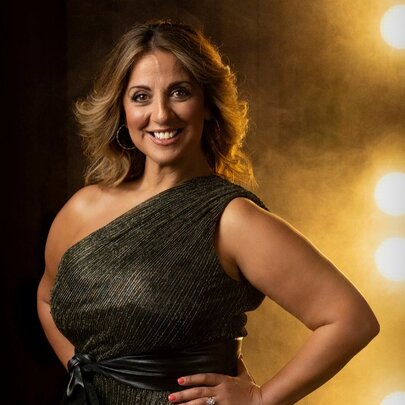 Portrait of Australian musical theatre star Silvie Paladino as she stands with her hands on her hips in a formal gown, in front of bright stage lighting.