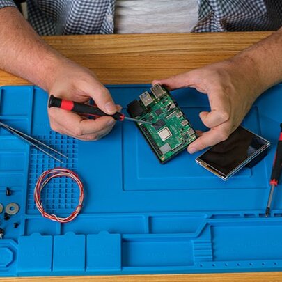 A pair of hands using a small screwdriver to work on an electronic circuit board, with other tools and wire nearby, all resting on a blue worksurface mat.