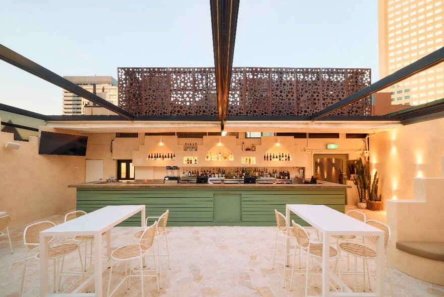 The open air rooftop bar at Hotel Nacional, with long white tables and chairs in a tiled space, a green coloured bar and soft wall lights.