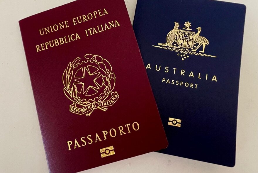 A photograph of a red Italian passport overlapping a blue Australian passport on a cream-coloured background.
