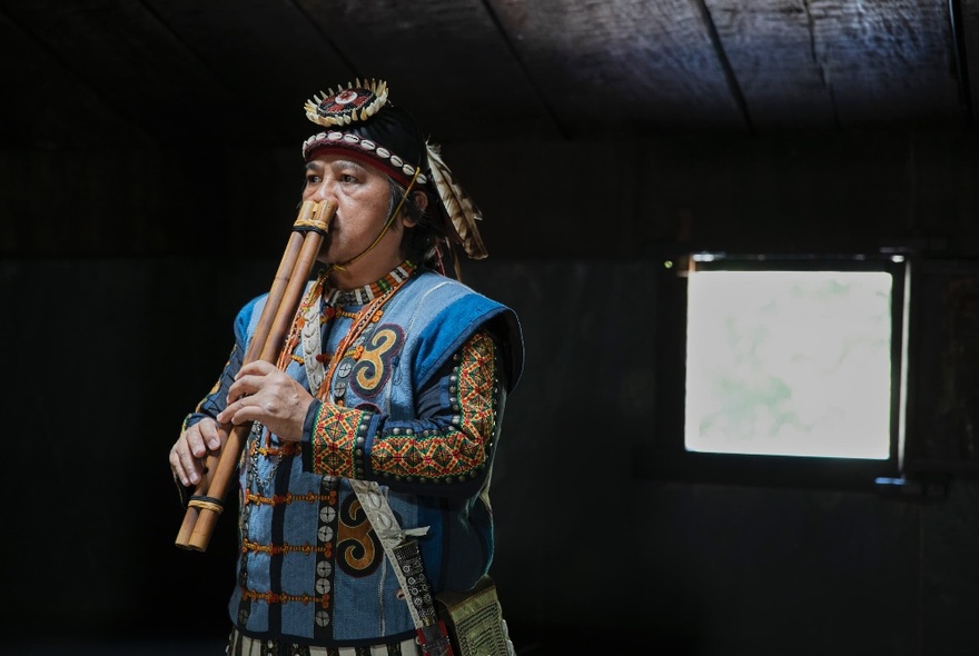 A person in a traditional Taiwanese costume playing a double-nosed flute with their nostrils, a square of light in the background. 