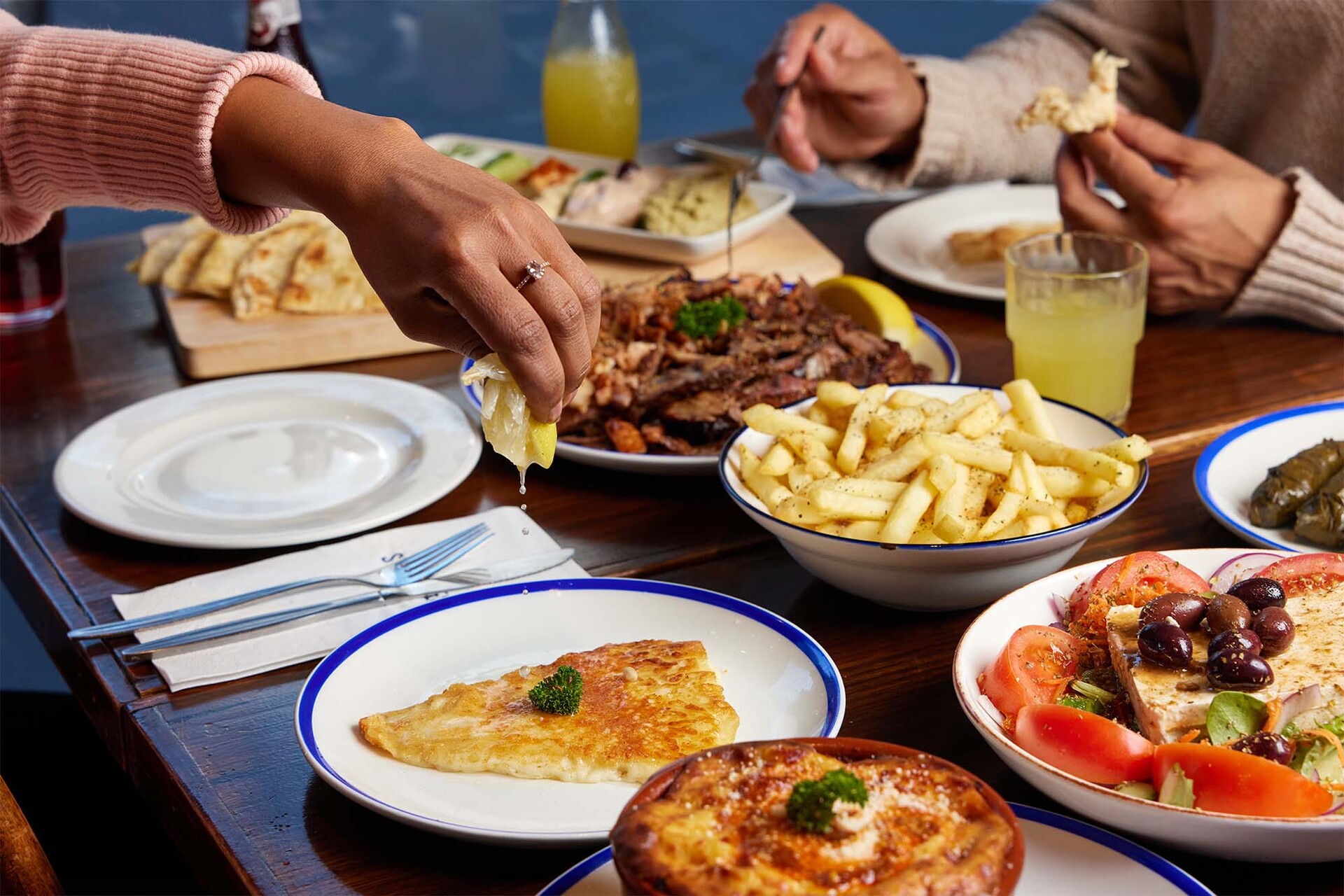 A table at a restaurant with plates of Greek food on it like lamb, salad and chips. A person is squeezing lemon onto the saganaki cheese.