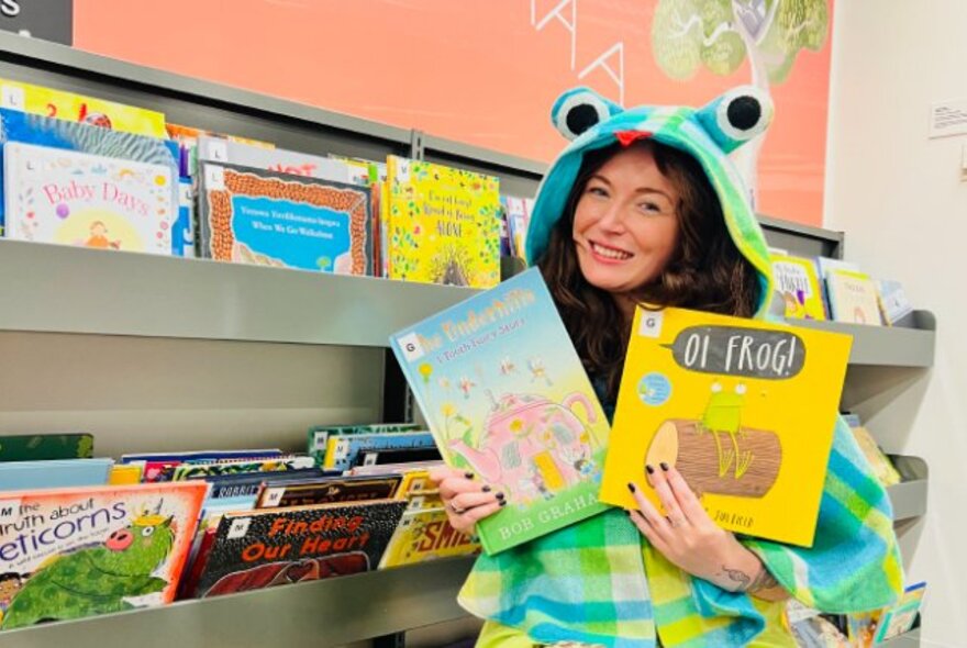 Woman wearing a frog coat with hood, holding books next to a bookshelf.