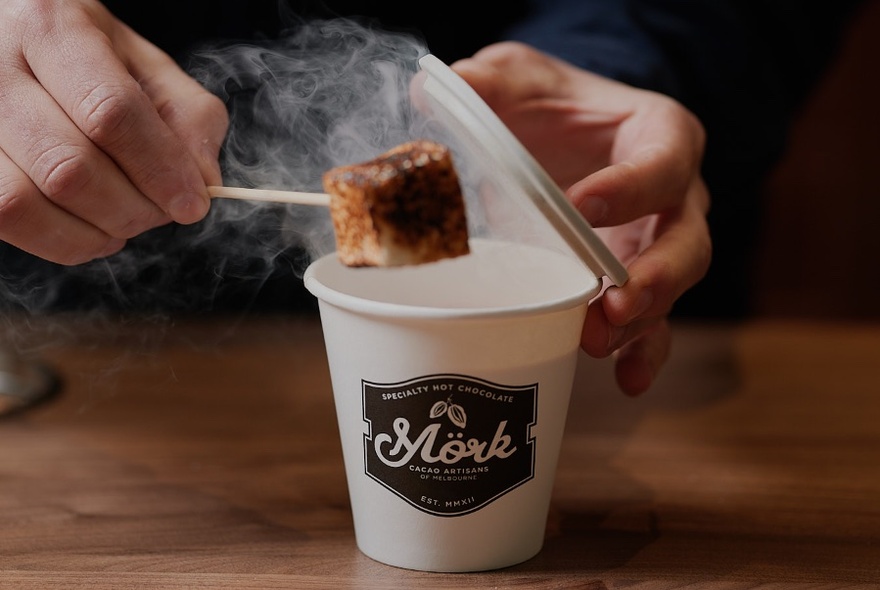 Takeaway coffee cup with left hand removing lid, right hand holding marshmallow over steam arising from cup.