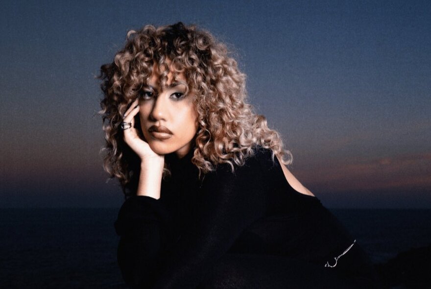 Performer Becca Hatch, her chin on her hand, with long curly hair, wearing black.
