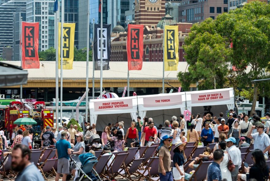 An outdoor exhibition with lots of people in Federation Square, KHT flags on poles. 