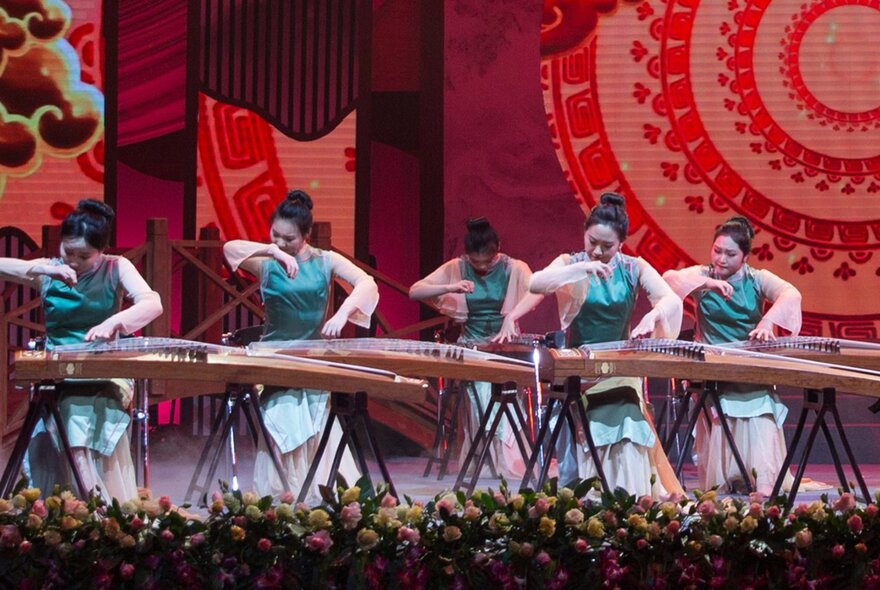 Chinese musicians playing a traditional instrument on stage, floral border in front of them and a red circular motif behind.