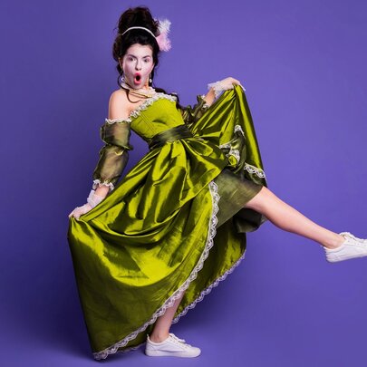 A person modeling a bright green, historical-style dress paired with modern white sneakers against a purple background.
