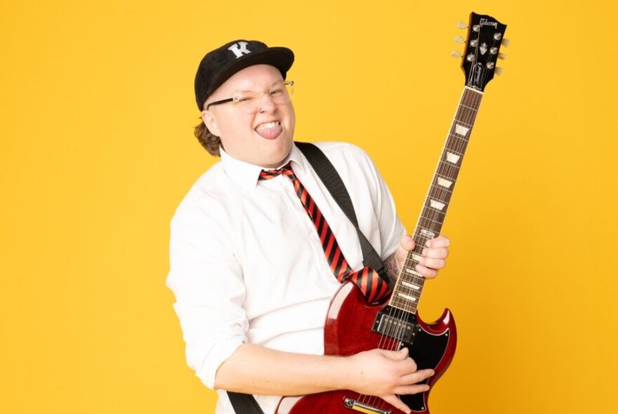 Comedian, Kaileb Hyland, wearing a white shirt, school tie, baseball cap and glasses, playing a guitar in front of a yellow background.