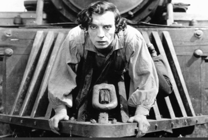 Black and white still from the 1920s film, The General, showing a young man riding on the front of a steam train.